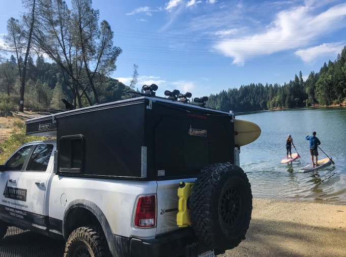 2 people paddle board while their pop-up camper sits on the shoreline.